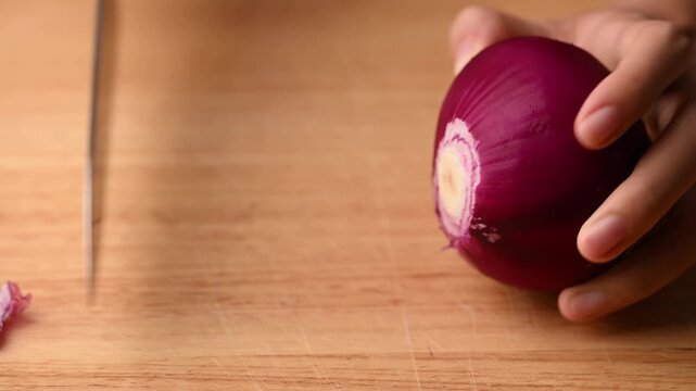 Close-up of woman's hands cutting a red onion in half with a knife on a wooden board, preparing ingredients for cooking