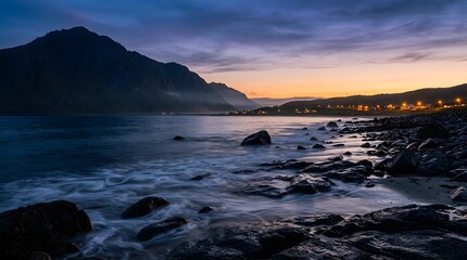Obraz premium Long exposure landscape of a rocky coast at dusk with mountains, ocean waves, and distant village lights under twilight sky