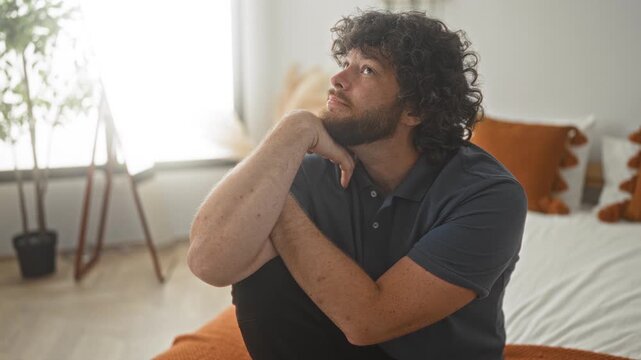 Man with hand on chin seated on bed in studio, young bearded man with curly hair wearing a navy polo, looking upward toward window in a softly lit bedroom scene; contemplation.
