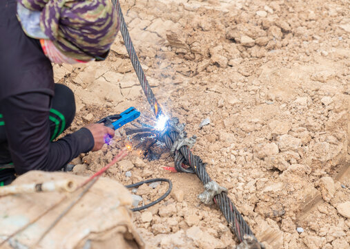 Construction worker using an arc welding torch to cut a heavy-duty steel wire rope cable on a dirt work site.
