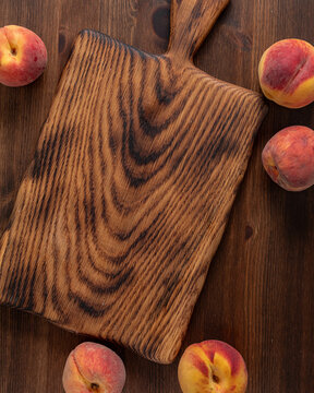 A wooden cutting board against a dark wooden surface and peaches all around.