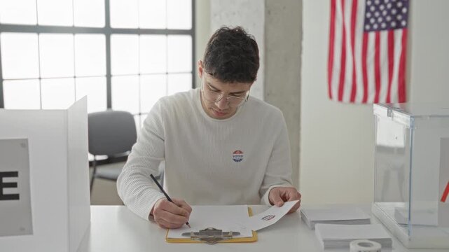 Man holding ballot and pen at a polling station table in building with transparent ballot box and us flag visible, wearing voted sticker; civic pride.