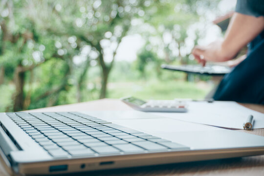 Laptop computer keyboard on wooden desk with calculator, document, and blurred person working in background with green nature trees view, concept of remote work and business office setup.