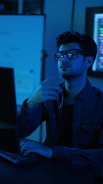 Young male programmer working late at night in a dark room illuminated by blue screen light. The focused IT professional types on a computer, coding and analyzing data with a thoughtful expression.