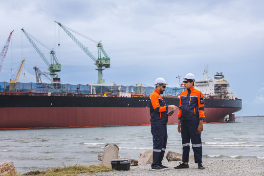 Maritime engineers discussing project at shipyard with cargo ship background, Port workers in safety gear reviewing documents at commercial harbor
