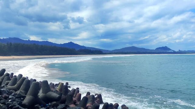 Ocean Waves Crashing Against a Rocky Barrier on a Serene Beach with Lush Green Mountains Under a Cloudy Blue Sky