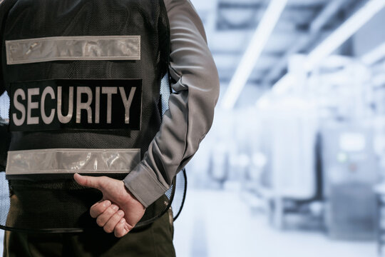 Close view of a security guard standing with hands behind back in a bright modern hallway, wearing a black vest with a security label.
