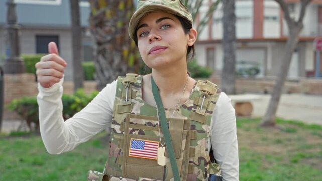 Woman soldier gives thumbs up wearing camouflage vest, cap and dogtags with usa flag patch while standing near building and park; pride duty.