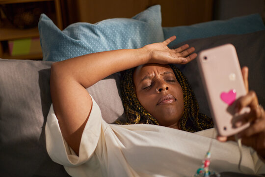 Tired African American young adult woman lying on sofa holding smartphone with hand on forehead, representing digital fatigue and suitable for mental health campaigns