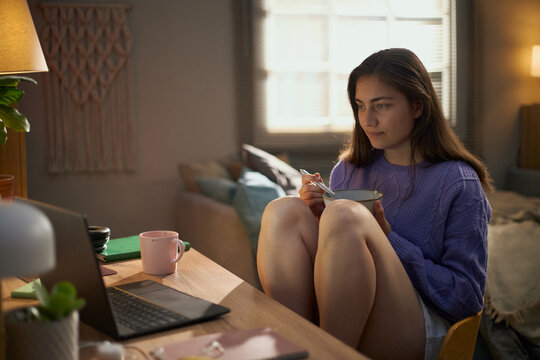 Young adult Caucasian woman eating breakfast while looking at laptop screen in cozy bedroom, representing remote work lifestyle and suitable for digital wellness campaigns