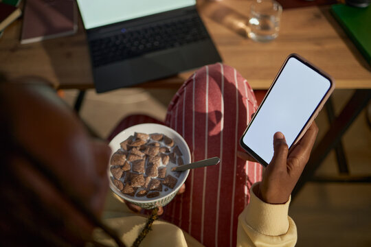 African American young adult woman holding smartphone with blank screen and eating breakfast cereal, representing remote work routine, suitable for mobile app marketing, high angle view