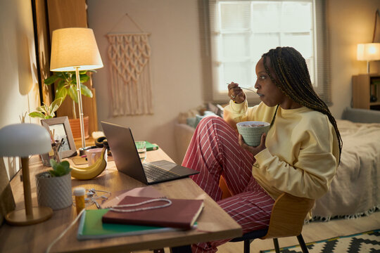 African American young adult woman eating breakfast while looking at laptop screen in bedroom, representing remote learning or work and morning routine, suitable for lifestyle marketing