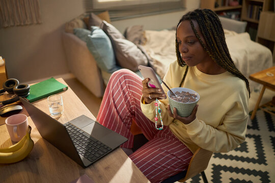 Young adult African American woman using smartphone while eating breakfast at desk with laptop, representing morning routine and remote work, ideal for lifestyle marketing