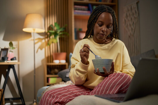 Young adult African American woman eating meal from bowl while watching laptop screen at home, representing remote lifestyle and digital entertainment, perfect for streaming service ads