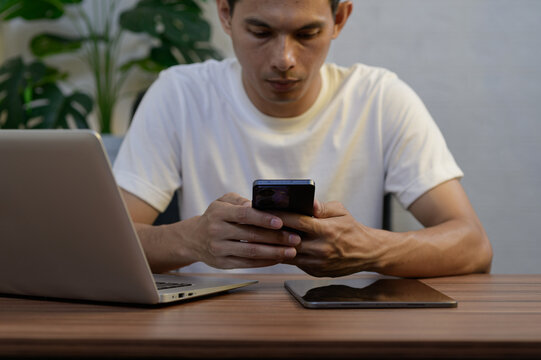 Young Man Using Smartphone While Working on Laptop at Desk