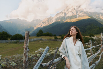 A young woman in a light poncho stands beside a wooden fence against a vast mountain landscape, radiating calm and curiosity in the outdoor scene. © SHOTPRIME STUDIO