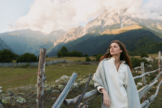 A young woman in a light poncho stands beside a wooden fence against a vast mountain landscape, radiating calm and curiosity in the outdoor scene.