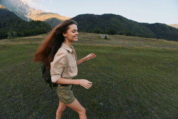 Naklejka premium Smiling woman with backpack hiking through a grassy field under mountains. Outdoors lifestyle and nature scene highlight adventure, travel, and carefree mood.