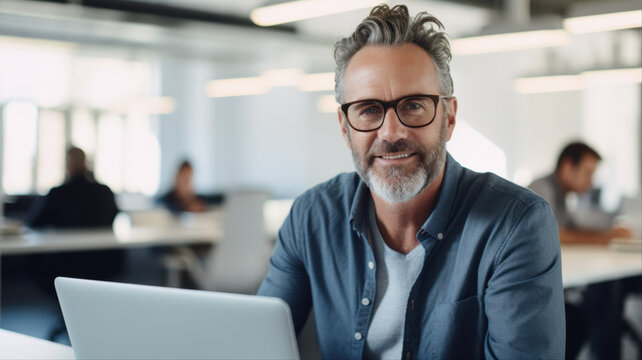 Confident Businessman in Modern Office: A distinguished businessman with salt-and-pepper hair and glasses smiles confidently at the camera, seated at his desk with a laptop in a bright.