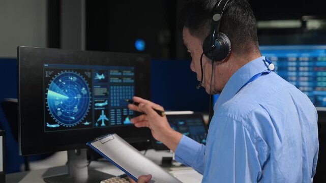Asian operator in headset typing and pointing at high-tech radar screens, managing telemetry data in modern control room.