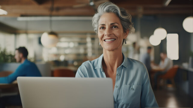 Confident Professional at Work: An experienced businesswoman with stylish gray hair smiles warmly while working on her laptop in a modern office setting.