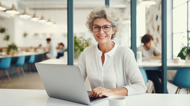 A Senior Professional Working: A confident and experienced senior woman with glasses sits at a desk, smiling at the camera while working on her laptop in a modern office environment.