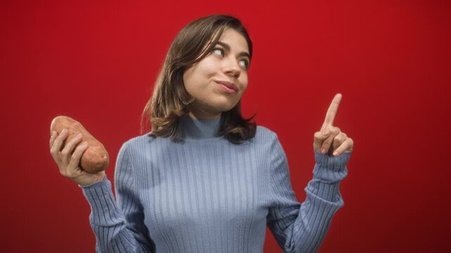 Woman holding sweet potato and pointing finger in studio, wearing blue turtleneck sweater with a sly smirk while looking upward; playful curiosity.