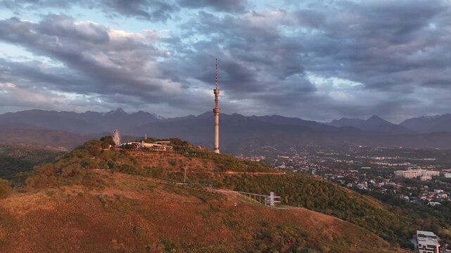 The famous TV tower on Kok Tobe Mountain in the Kazakh city of Almaty, set against a mountain range on a cloudy summer evening.