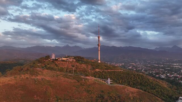 The famous TV tower on Kok Tobe Mountain in the Kazakh city of Almaty, set against a mountain range on a cloudy summer evening.