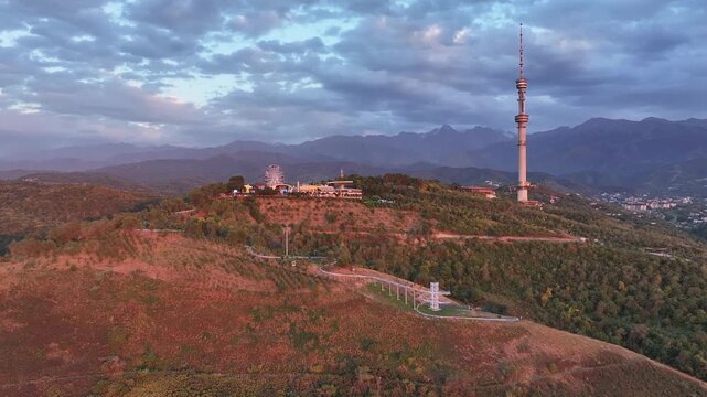 The famous TV tower on Kok Tobe Mountain in the Kazakh city of Almaty, set against a mountain range on a cloudy summer evening.