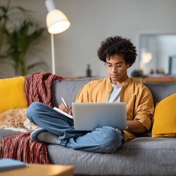Young man wearing glasses working with laptop and notebook on a comfortable couch
