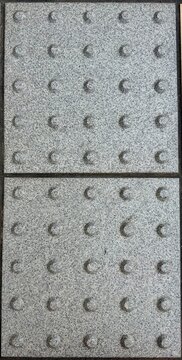 Close-up of gray tactile warning paving with raised dots designed to assist visually impaired pedestrians, showing a textured stone surface and repeating geometric pattern.
