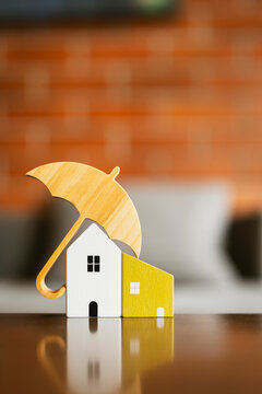 Home insurance and property protection concept with wooden umbrella covering miniature houses on reflective table with brick background for real estate mortgage, safety and risk coverage.