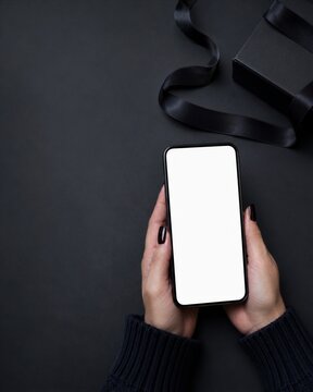 Woman hands holding smartphone with blank white screen mockup next to black gift box with ribbon on dark background. Online shopping and black friday sale concept with copy space