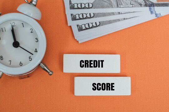 "CREDIT SCORE" text on wooden blocks with a white alarm clock, and US dollar bills on a coloured background. Represents financial deadlines, loan approvals, interest rates, and the urgency of managing