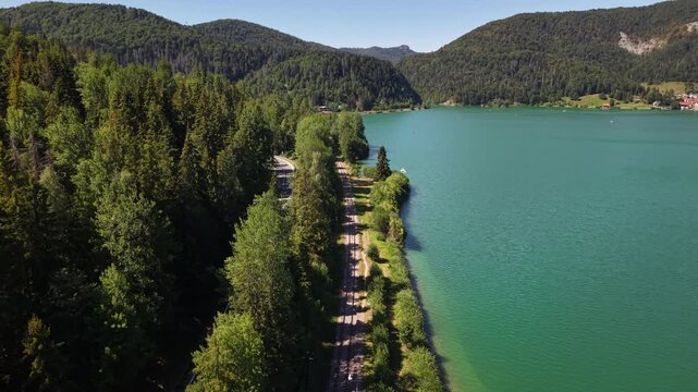 An aerial view captures a tranquil turquoise alpine lake, bordered by lush green forests, mountains, railway tracks, and a parallel road.