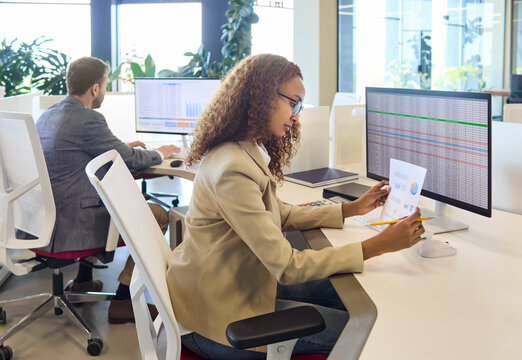 Businesswoman analyst reviewing financial spreadsheet at computer. In a bright office she checks charts on a monitor and report while coordinating budgets. Teamwork and data accuracy focus.