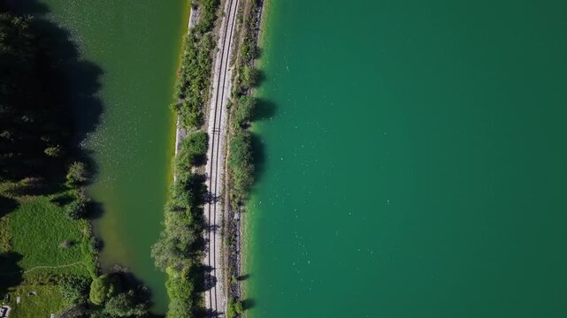 An aerial perspective shows train tracks bordering a vibrant, green lake surrounded by dense forest and grassy hills.