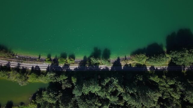 An aerial view shows train tracks running alongside a vibrant green lake, bordered by dense forest and trees.