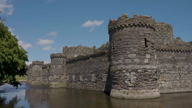 View of Beaumaris Castle with its extensive moat clearly visible in the foreground, showing calm water, weathered stone walls and towers surrounded by rich green grass and natural countryside.