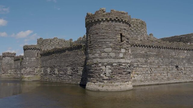 View of Beaumaris Castle and its surrounding moat, with calm water in the foreground leading toward the weathered medieval walls, towers and green grassy grounds of this historic Welsh landmark.