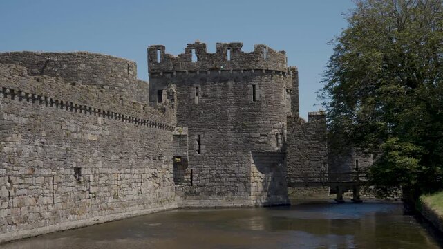 View of the moat surrounding Beaumaris Castle, with ancient stone walls rising behind the water and green vegetation covering the surrounding grounds, emphasising the castle medieval defensive design.