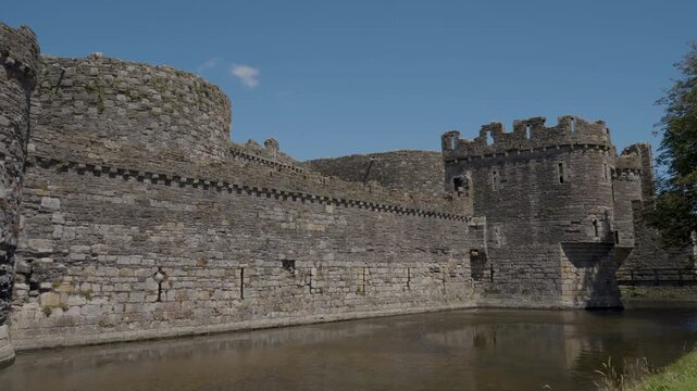View of Beaumaris Castle and its moat from the surrounding grounds, showing the water-filled defensive channel, imposing stone towers and extensive medieval walls within a lush green landscape.