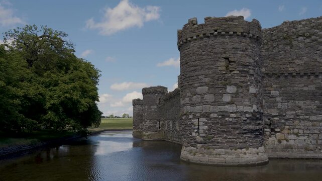 View across the moat toward Beaumaris Castle, capturing the imposing stone walls and towers rising behind the water, green vegetation creating a peaceful and atmospheric historic landscape.