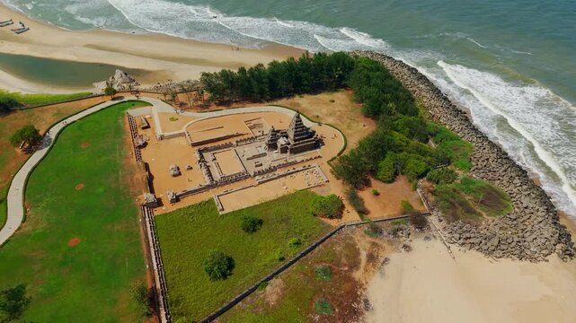 Cinematic aerial view of Shore Temple in Mahabalipuram, Tamil Nadu, India, set beside the Bay of Bengal coastline, sandy beach, waves, boats, green grounds and historic temple architecture.