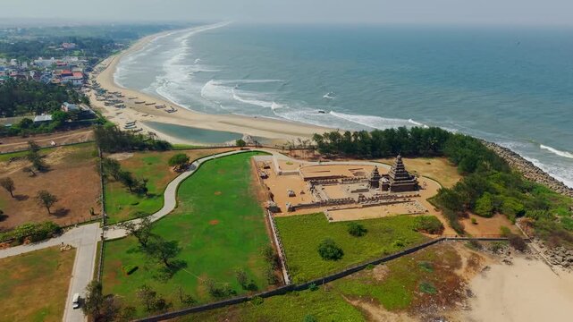 Cinematic aerial view of Shore Temple in Mahabalipuram, Tamil Nadu, India, set beside the Bay of Bengal coastline, sandy beach, waves, boats, green grounds and historic temple architecture.