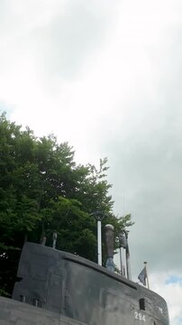 A low angle shot of a decommissioned submarine tower and periscope equipment at the Polish Naval Museum in Gdynia, set against green trees and a cloudy sky.
