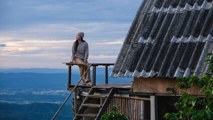 Fototapeta premium A woman sitting on wooden balcony, looking at mountain views on cloudy morning