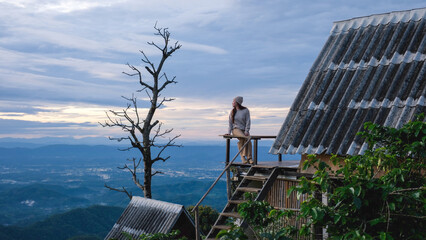 Fototapeta premium A woman sitting on wooden balcony, looking at mountain views on cloudy morning