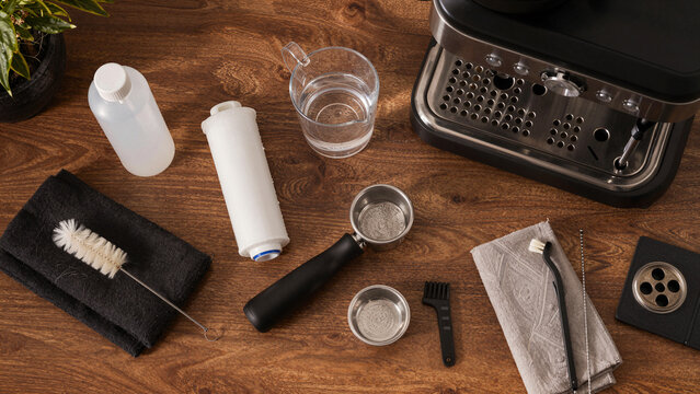 A variety of tools and supplies are laid out on a wooden table. Items include a water container, a cleaning brush, and machine components. This setup is for cleaning a coffee machine